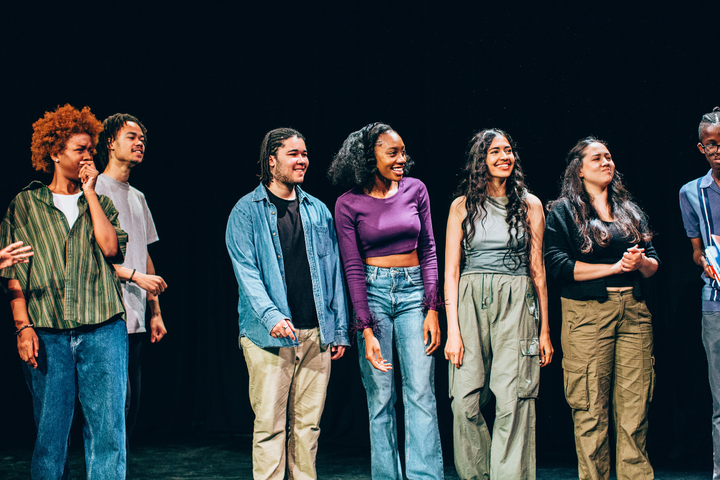 Originate students smile in a circle looking stage left, acting on stage.