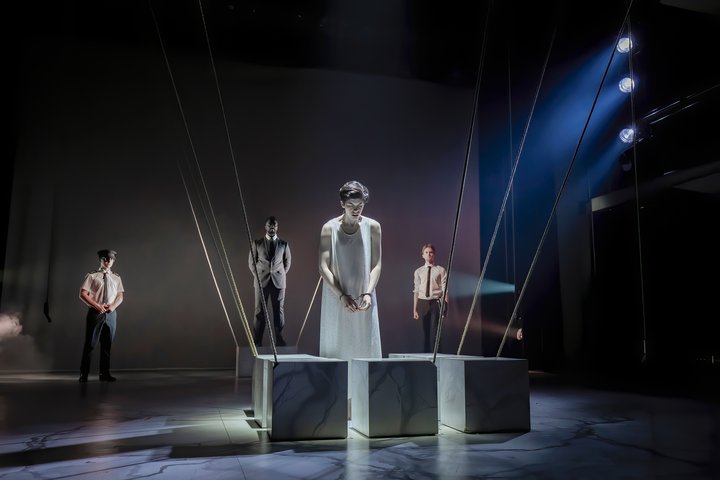 Four actors appear on a dramatically lit stage next to floor-length ropes tethered to the ceiling and behind three marble-effect squares.