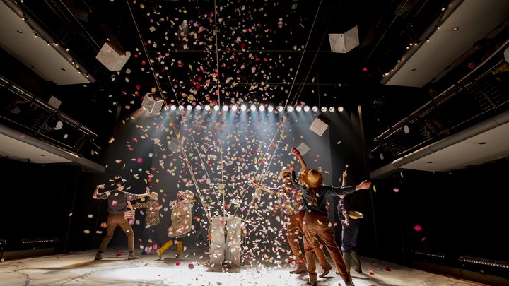 A group of actors look celebratory, moving around red and white confetti in warm lighting on stage.
