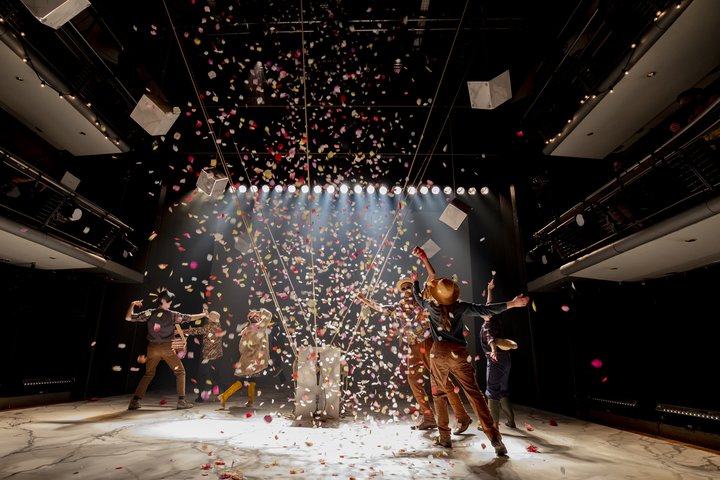A group of actors look celebratory, moving around red and white confetti in warm lighting on stage.