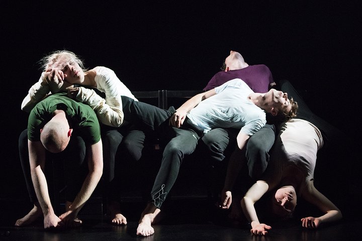Figures on a dark background lit in bright white light lying as if asleep piled up on stage