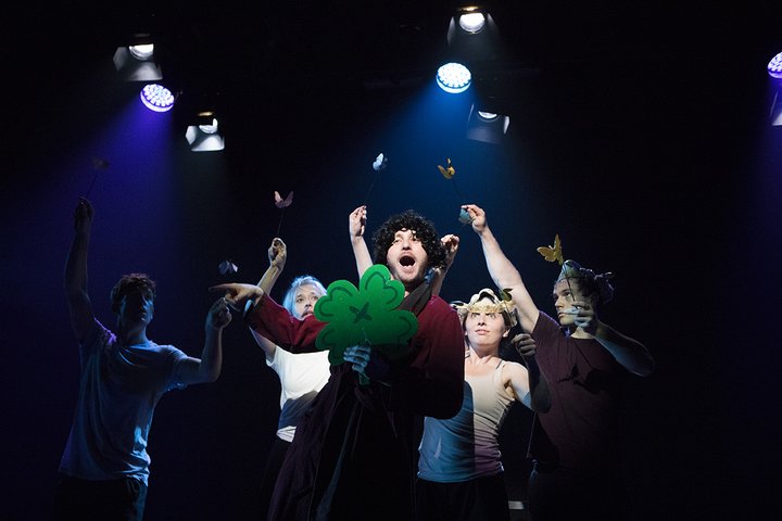 Figures grouped together in silly costumes around a shamrock looming out of a dark stage