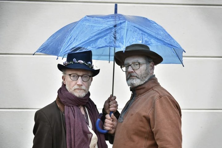 Jeremy Stockwell and Chris Larner stand under a shared blue umbrella in a promo shot for A Sockful of Custard