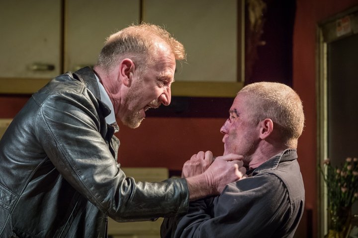 Adam Gillen (right), with cropped hair and bruises and cuts on his face, is seized by the collar angrily by another actor, in a scene from Killer Joe