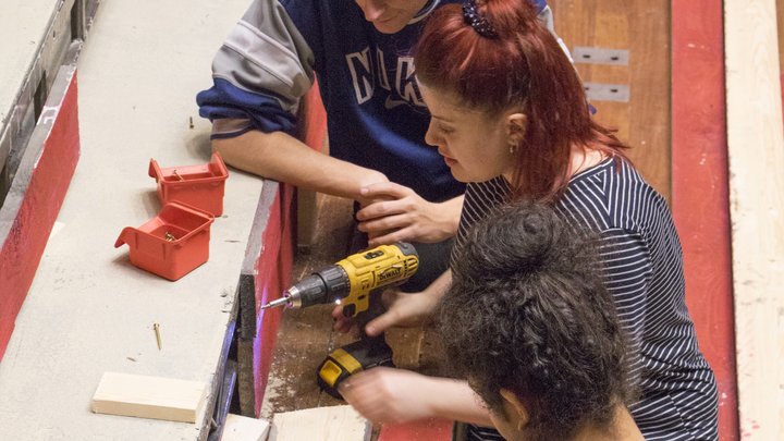 Three students work on a theatre set on the front of the stage. Bethany is in the centre of the three, drilling into a wooded panel.