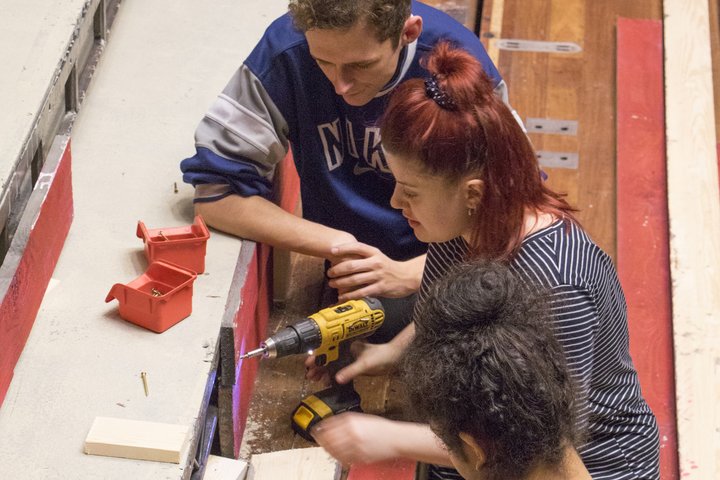 Three students work on a theatre set on the front of the stage. Bethany is in the centre of the three, drilling into a wooded panel.