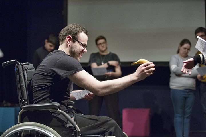 A young man who is a wheelchair user dressed in black disabled acting and holding a banana in front of himself laughing.
