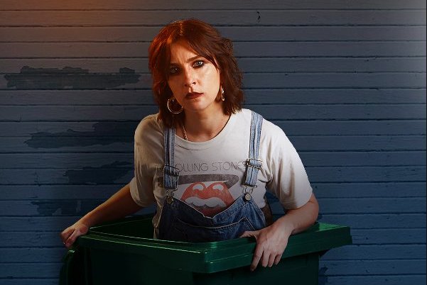 Karen Cogan dressed in dungarees stands inside wheelie bin in a promo shot for her show Drip Feed