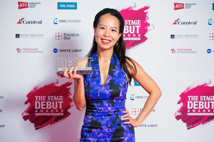 RADA MA Theatre Lab graduate Elisabeth stands in front of The Stage branded wall wearing bright blue dress with Debut Award in hand