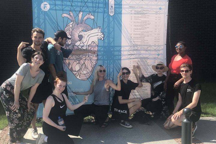Group of actors stand and sit in front of blue poster and white posters, pointing at listings of performances in Gdansk