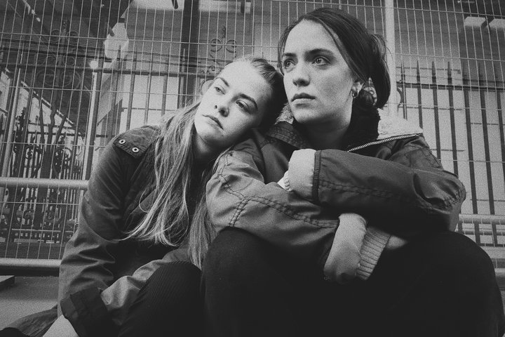 In a black white photo, two RADA students and cast members of Cathy sit in front of builders' railings on a city street