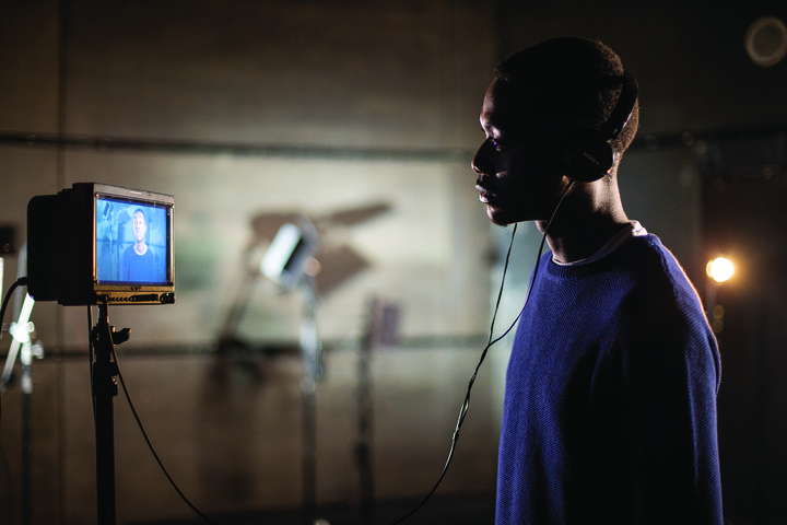 © Chris Vile A RADA acting student stands on the right of the frame in headphones, watching footage of himself on a screen on the left of the frame