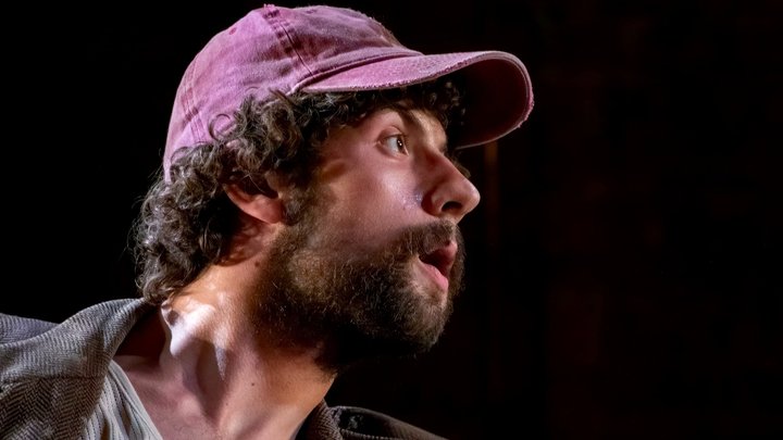 Close up of Keaton's side profile as he looks right in dramatic lighting while wearing a cap.