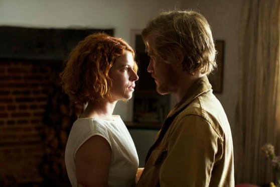 Actress Jessie Buckley stares into the face of co-star Johnny Flynn in a domestic room. The room is in shadow but light falls on her face.