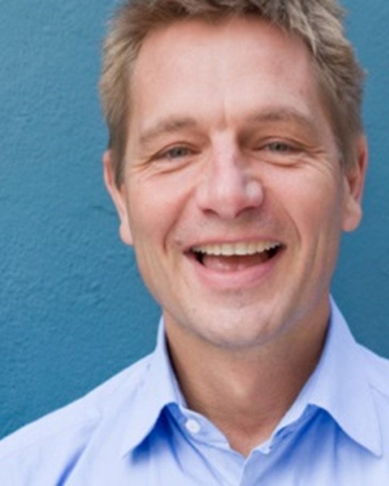 Head and shoulders of John Tucker smiling and looking directly at the camera. John stands in front of a blue background.
