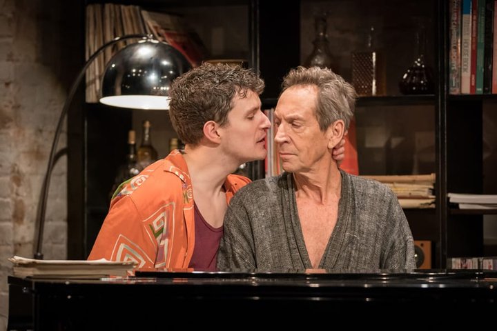 Two actors, one younger and one older with grey hair, sit side by side at a grand piano. The younger man rests his hand behind the other's neck.