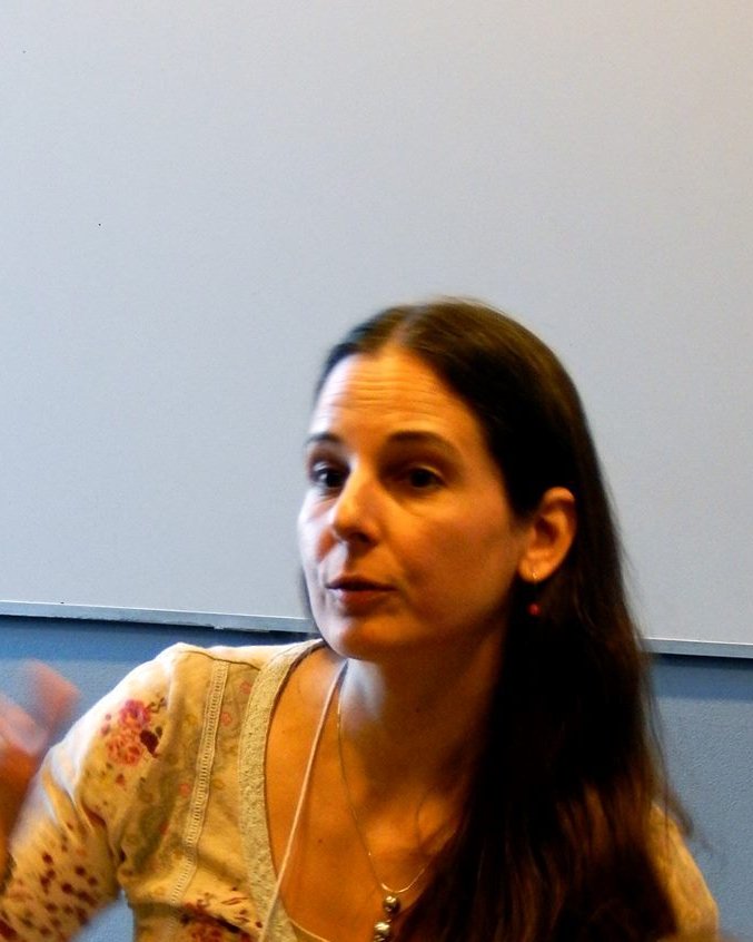 head and shoulders of woman with long dark hair gestures towards camera