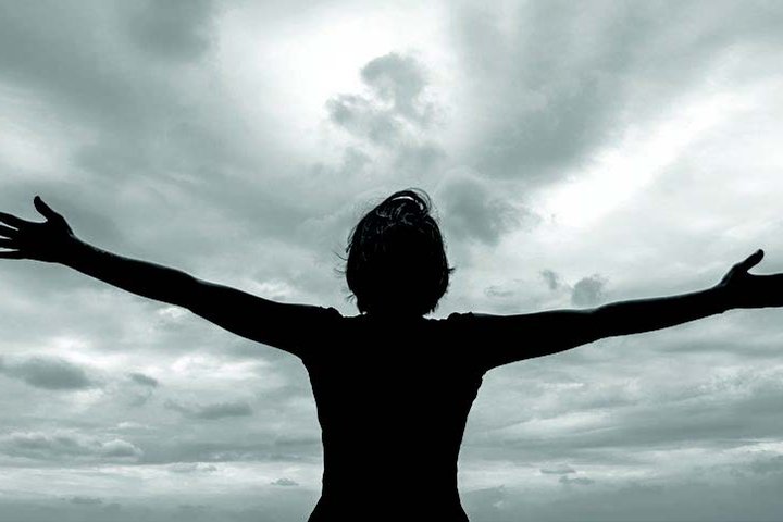 A woman stands, back to the camera, looking up at a wide open sky with arms outstretched