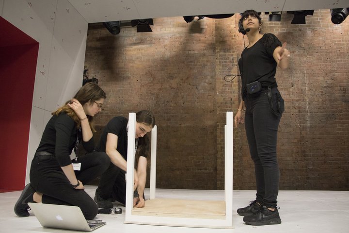 On a lit stage in front of an exposed brick wall, two student stage managers fix an upturned table while a third looks up to the tech box.