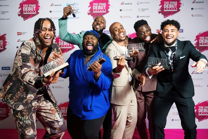 A group of men including BA acting graduate Nnabiko Ejimofor laughing and posing in front of The Stage branded wall holding Debut awards