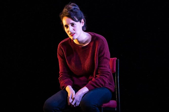 In a dark red jumper with hair tied back, Phoebe Waller Bride sits on a dark stage with a spotlight on her during her West End performance of Fleabag