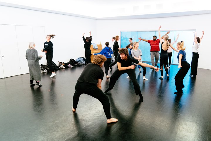 A mixed group of young actors in rehearsal clothing in a studio. Their arms are up and the energy is high for a movement class.