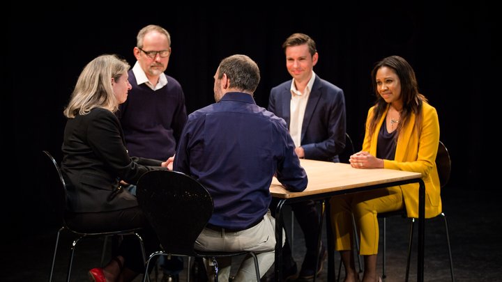 A RADA Business tutor and participants sit around a board table on a stage. The auditorium is empty and the spotlights are shining on them.