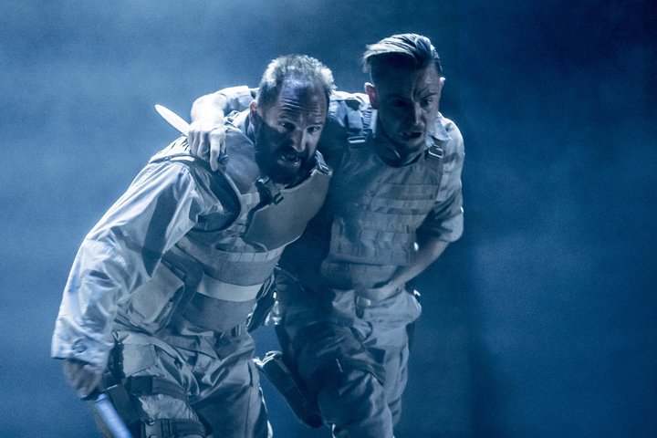 Ralph Fiennes and Alexander Cobb, dressed in contemporary army uniform and carrying knives, hold each other up in a scene from Antony and Cleopatra