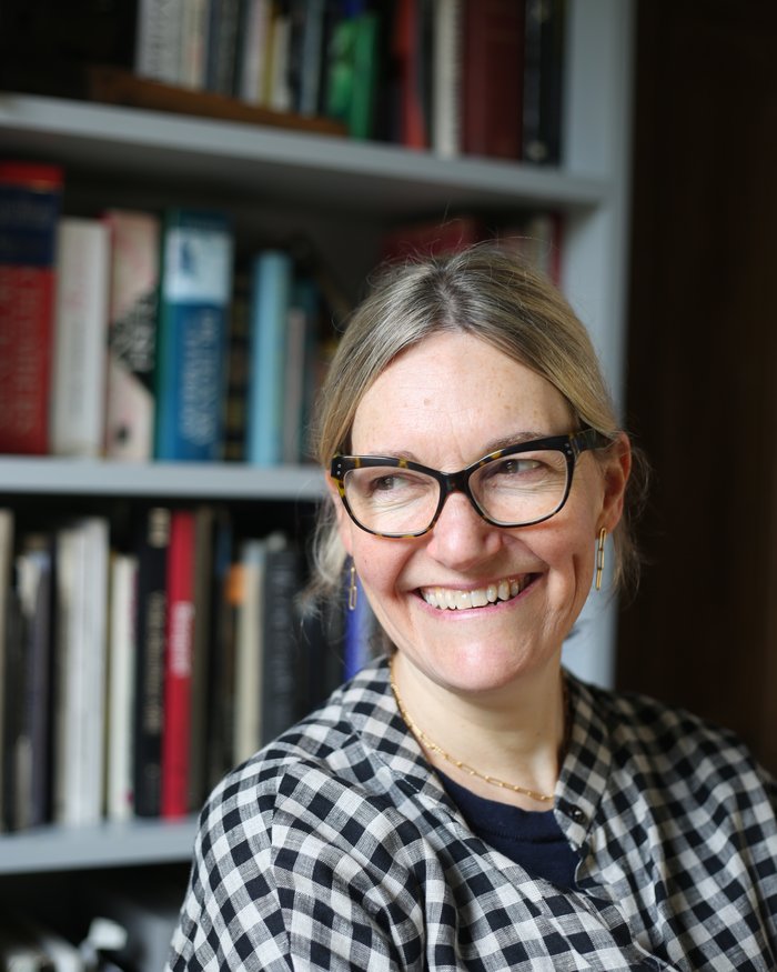 Tanya Ronder sits in front of a bookcase, smiling while glancing to her right wearing dark tortoiseshell glasses.
