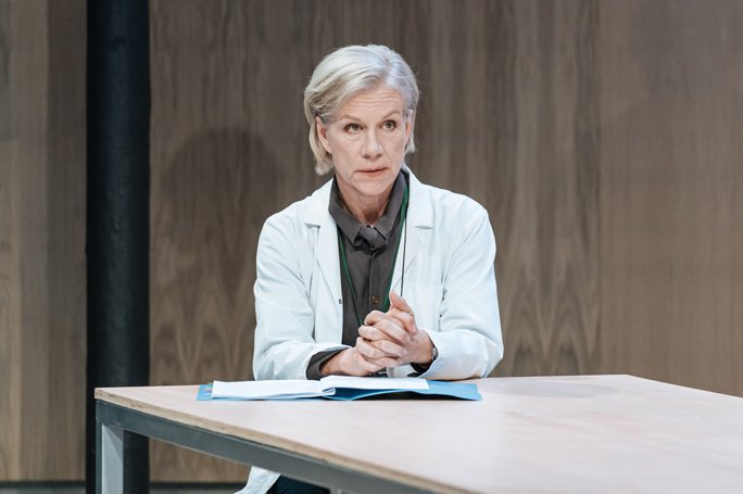Juliet Stevenson sits at a table, hands clasped with a serious expression, during her performance in The Doctor at the Almeida Theatre, London