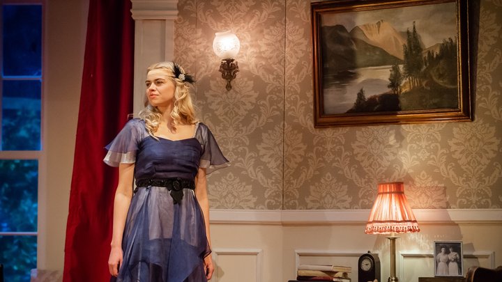 Mila Carter stands in a dress with chiffon overlay gazing across a room with a confident look