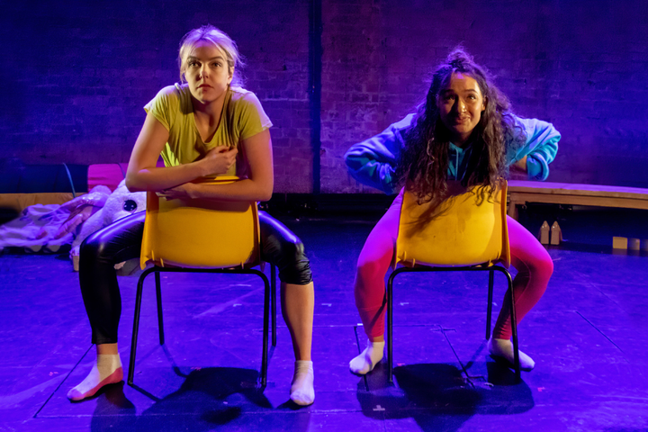 Two actors sit on yellow chairs in dramatic blue lighting with nervous expressions.
