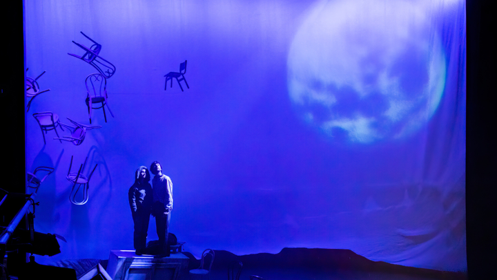 Two actors stand on stage in dramatic lighting looking upwards. The set features chairs floating in mid-air and a large image of the moon.