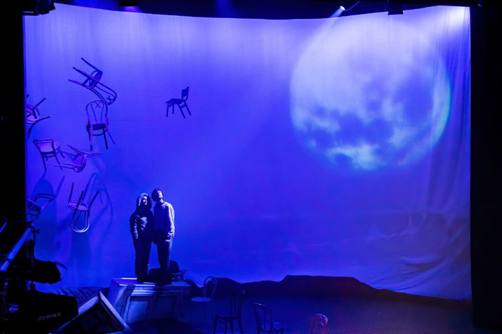 Two actors stand on stage in dramatic lighting looking upwards. The set features chairs floating in mid-air and a large image of the moon.