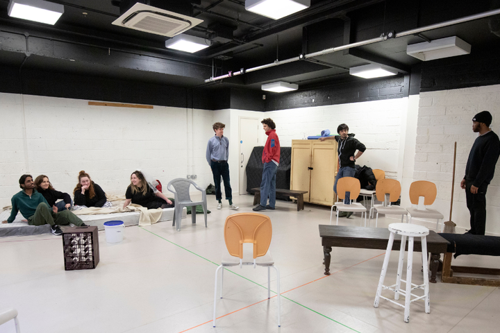 Eight actors sit and stand in the rehearsal room for Wild Cherries. They are surrounded by chairs and furniture making up the set.