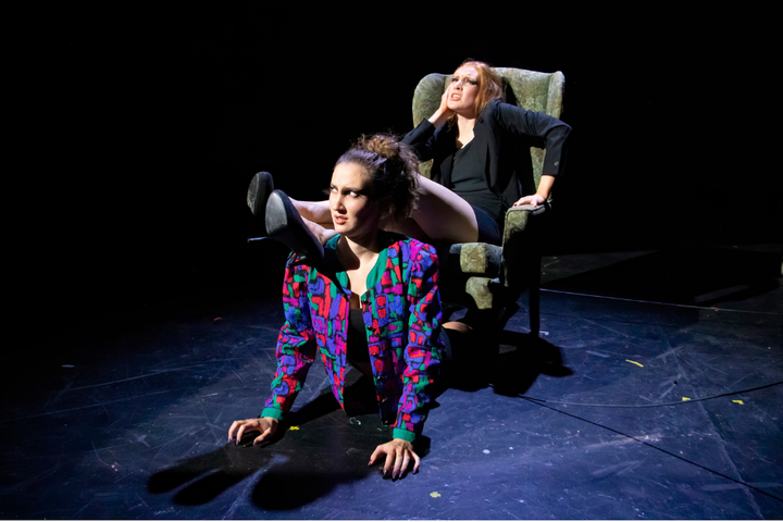Two actors perform on stage in bright, dramatic lighting. One actor sits in a suit and the other lies on the floor in a brightly coloured jacket.