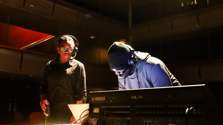 A young technician in a dark beanie hat works at a sound desk. Another young technician looks over the work with a headset on.