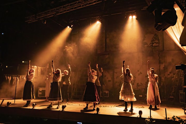 A group of actors look defiant on stage under sepia lighting with fists raised in the air.
