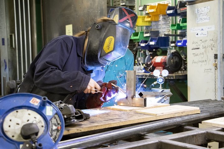 In a construction workshop, a student wearing a face shield welds a metal joint, causing blue sparks to fly
