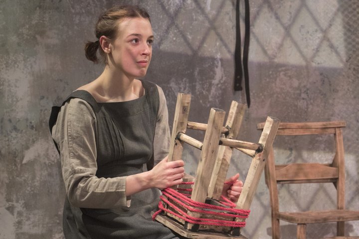 Actress Lucie Sword sits on stage in RADA's GBS Theatre holding wooden stool wrapped in red thread in a production of The House of Bernarda Alba