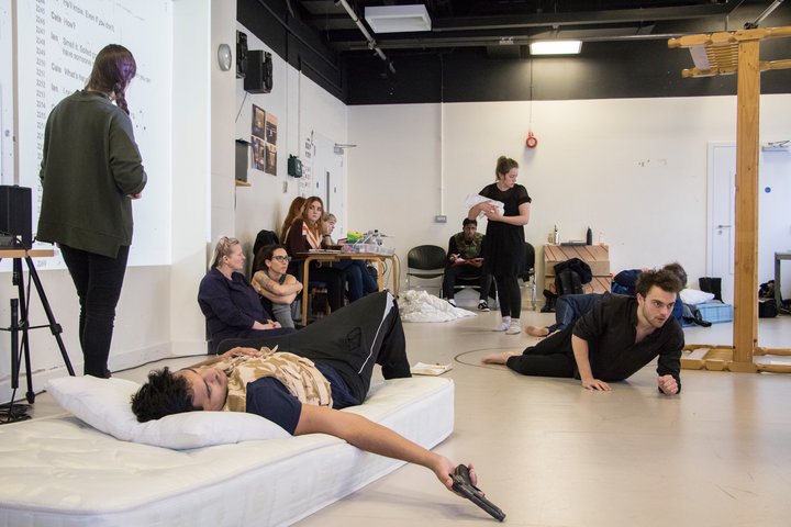 A group of people including actors, directors are working on a scene. There is a man lying on a bed in the middle lazily holding a gun.