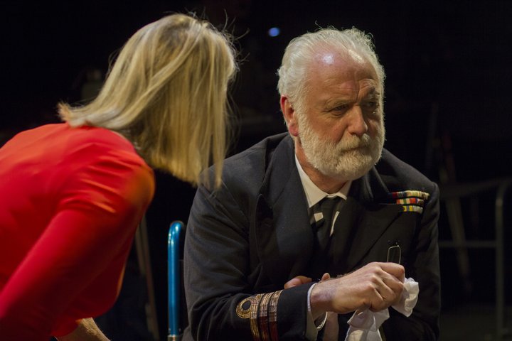 A bearded older man in navy-style uniform sits with hands folded looking into the distance. A blonde women in red stands looking into his face.