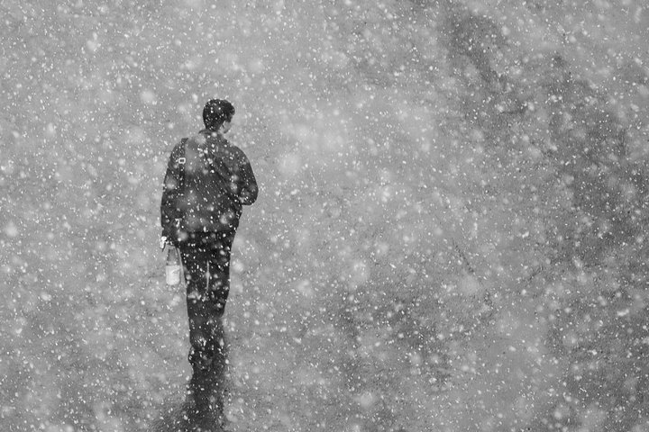 In a black & white image, a man walks away from the camera carrying a bottle of champagne through a blizzard in an empty landscape