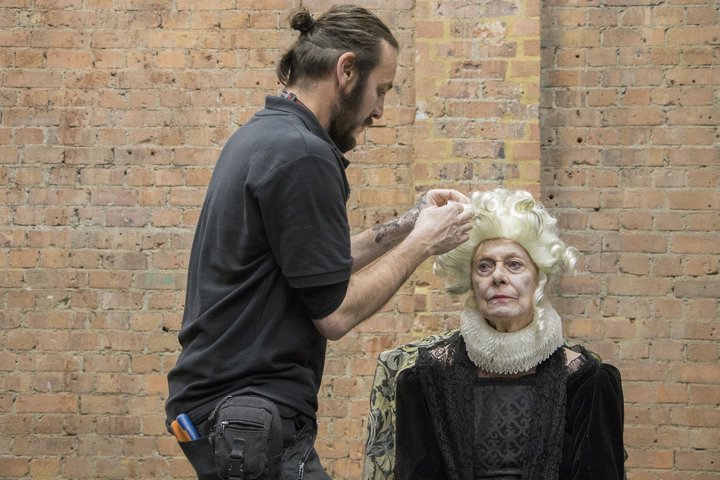 An older actress is seated in front of a brick wall in a black lace Tudor period costume, while a stage hand adjusts her blonde, curled wig