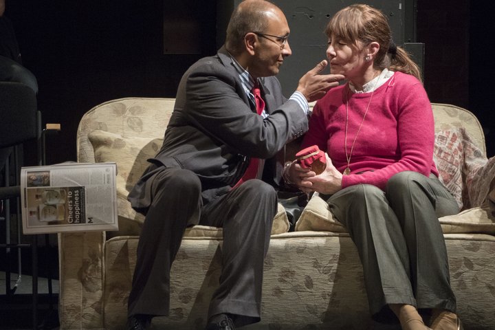 A male and female actor from the Elders Company sit looking at each other on a cream sofa. The man's hand is raised affectionately to the woman'sface.