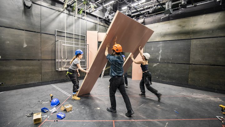In the Gielgud Theatre at RADA, students in hard hats raise a flat wall into position as part of a set.