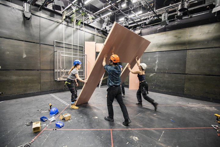In the Gielgud Theatre at RADA, students in hard hats raise a flat wall into position as part of a set.