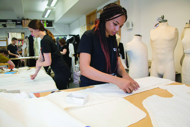 A girl with long braids stands at a workshop table cutting patterns for a costume