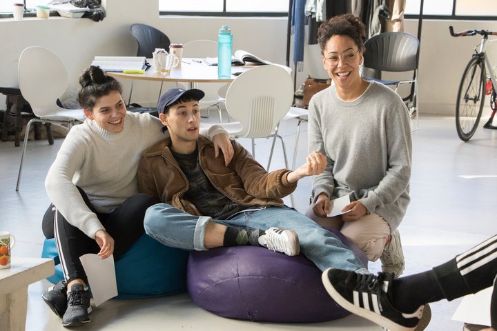 Three students sit on the floor. In the centre, a male student in cap and jeans reclines on a purple bean bag gesturing to someone off camera.