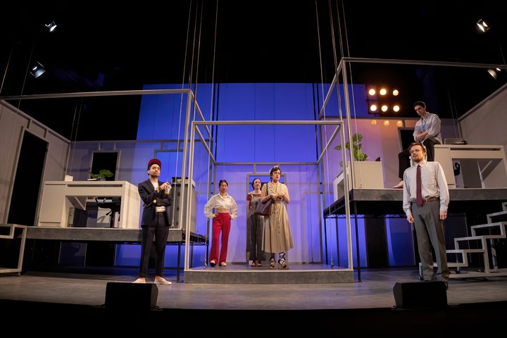 Production image from Gloria performed at RADA in 2023. Six actors stand on a multi-level stage which has been designed as an office.